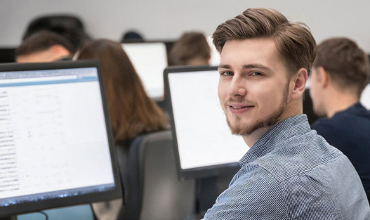 Leende ung man i ett klassrum med studenter tittar på datorskärmar